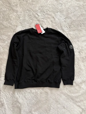 Cp company tröja - Stilren svart sweatshirt med rund halsringning och ribbade muddar. Tröjan har en diskret patch på ena ärmen och en liten ficka med dragkedja. Perfekt för en avslappnad och trendig look.