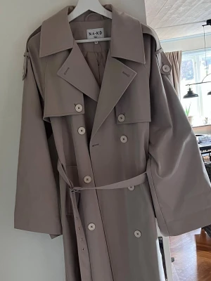 Beige trenchcoat från NA-KD - Stilren beige trenchcoat från NA-KD med klassisk dubbelknäppning, bred krage och bälte i midjan. Jackan har långa ärmar och axelklaffar för en trendig look. Perfekt för lager-på-lager under vår och höst.