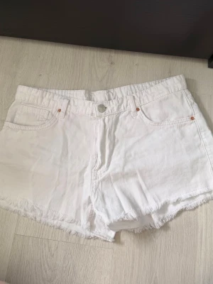 Vita jeans shorts  - Säljer ett par vita jeansshorts med fransig kant nertill. Klassisk femficksmodell med knapp och dragkedja framtill. Perfekta till sommaren och lätta att matcha med olika toppar. Gjorda i bomull för en skön och luftig känsla.