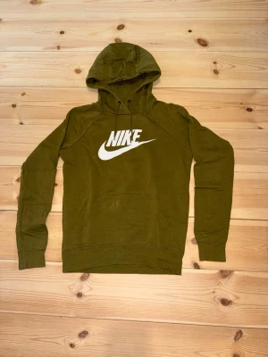 Olivgrön Nike hoodie med logga - Snygg olivgrön hoodie från Nike med stor vit logga på bröstet. Hoodien har huva med dragsko och en klassisk känguruficka framtill. Tillverkad i mjukt material och har ribbade muddar vid ärmar och nederkant. Perfekt för en avslappnad och sportig stil.