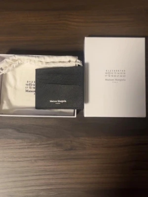 Maison Margiela plånbok  - Snyggt svart kortfodral från Maison Margiela i läder med strukturerad framsida och vita sömdetaljer på baksidan. Fodralet har flera kortfack och minimalistisk design med Maison Margiela-logga framtill. Perfekt för dig som gillar stilrena accessoarer.