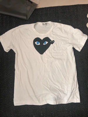 Comme des Garçons t-shirt - Säljer en vit t-shirt från Comme des Garçons Play med det ikoniska svarta hjärtat med blå ögon tryckt på bröstet. Klassisk rund hals och korta ärmar. Tillverkad i mjuk bomull för en skön känsla. Perfekt för dig som gillar streetwear och stilrena plagg.