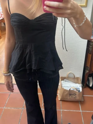 Svart volang topp med tunna band - Säljer en svart topp med tunna axelband och volang nedtill. Toppen har en söt rynkning över bysten och sitter figurnära upptill. Perfekt att styla med jeans eller kjol för en trendig look.