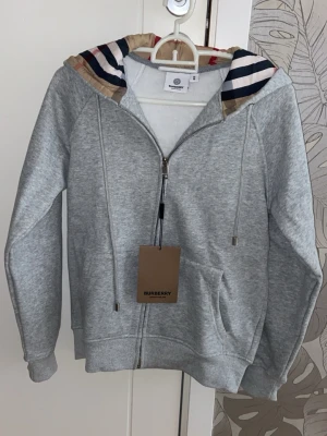 Burberry Hoodie - Snygg grå hoodie från Burberry med dragkedja och klassiskt rutigt mönster i huvan. Mjuk bomullsblandning, två fickor fram och justerbar huva med snören. Perfekt för en chill och stilren look.