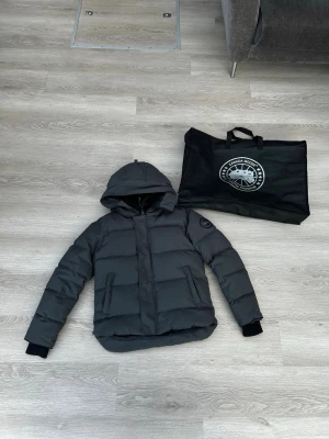 Svart dunjacka från Canada Goose - Säljer en svart dunjacka från Canada Goose med huva och två sidofickor. Jackan har en diskret logotyp på ärmen och muddar vid ärmsluten. Perfekt för kalla vinterdagar och riktigt snygg till streetwear-stilen.