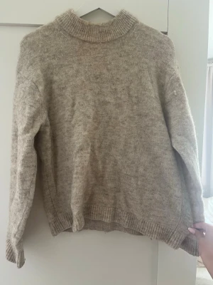 Beige stickad tröja med polokrage - Mysig beige stickad tröja med hög polokrage och långa ärmar. Tröjan har en avslappnad passform och ribbstickade muddar vid ärmslut och nederkant. Perfekt för kyliga dagar och enkel att matcha med olika outfits.