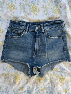 Blå jeansshorts med rå kant - Säljer ett par klassiska blå jeansshorts med hög midja och rå, fransig kant nedtill. Shortsen har fem fickor, bälteshällor och stängs med knapp och dragkedja.  