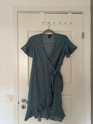 Blå prickig omlottklänning Vero Moda - Söt blå omlottklänning från Vero Moda med vita prickar och knyt i midjan. Klänningen har korta ärmar, volangkant nertill och v-ringad hals. Perfekt för varma dagar och ger en avslappnad men ändå trendig look. Använd ett fåtal gånger☺️