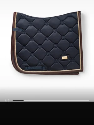 ES schabrak  - Snyggt mörkblått schabrak med quiltat mönster och guldfärgad kant. Har bruna detaljer runtom och en liten metallbricka på sidan. Perfekt till hästutrustning och ger en stilren look i stallet.