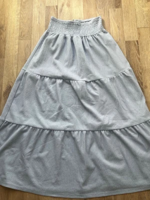 Ljusblå långkjol med smock - Säljer en ljusblå långkjol med bred smock i midjan och volangdetaljer. Kjolen har en mjuk och flowy känsla, perfekt för varmare dagar. Materialet känns som bomull eller polyester och färgen är diskret ljusblå.