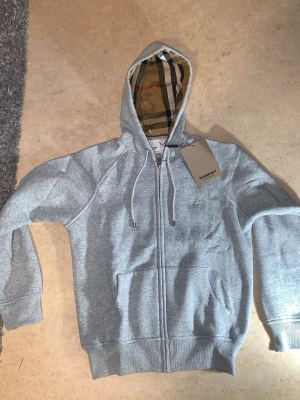 Burberry zip hoodie  - Snygg grå zip hoodie från Burberry med klassiskt rutigt foder i huvan och broderad logga på bröstet. Hoodien har dragsko i huvan, fickor framtill och ribbade muddar. Perfekt för en chill och stilren look.