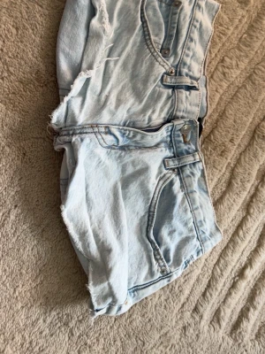 Ljusblå denimshorts med rå kant - Säljer ett par ljusblå denimshorts med råa, fransiga kanter och klassisk femficksdesign. Shortsen har knappgylf och är perfekta för varma dagar. Snygg och avslappnad stil som passar till det mesta.