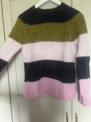 Randig stickad tröja - Säljer en stickad tröja med breda ränder i blått, olivgrönt och ljusrosa. Tröjan har rund hals, lång ärm och är gjord i ett mjukt, fluffigt material som ger en cozy vibe. Perfekt för dig som gillar färgkombinationer och oversized känsla.