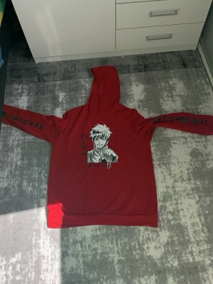 Röd anime hoodie med tryck - Cool röd hoodie med stor huva och anime-tryck på bröstet i svartvitt. Textdetaljer på båda ärmarna ger extra attityd. Perfekt för dig som gillar streetwear och manga-inspirerad stil. Mjuk och skön bomullskänsla.