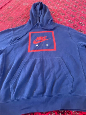 Blå Nike Air hoodie med röd logga - Säljer en blå hoodie från Nike Air med stor röd och vit logga på bröstet. Hoodien har huva med dragsko och en stor magficka framtill. Tillverkad i mjukt material som passar perfekt för chill dagar. Klassisk streetwear-look.