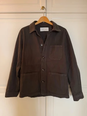 Mörkgrön overshirt från A Day's March - Stilren mörkgrön overshirt från A Day's March med tre stora utanpåfickor framtill och knäppning med fyra knappar. Overshirten har klassisk krage och raka ärmar. Tillverkad i ett kraftigare bomullstyg som ger en avslappnad men snygg look.