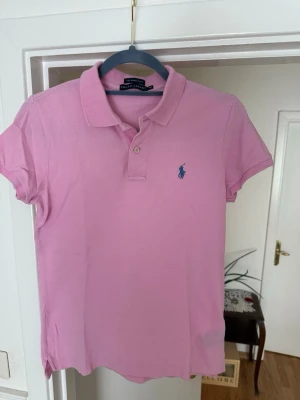 Rosa pikétröja från Ralph Lauren, figursydd  - Säljer en rosa pikétröja från Ralph Lauren i modellen The Skinny Polo. Tröjan har klassisk krage, korta ärmar och två knappar framtill. Den ikoniska loggan i blått är broderad på bröstet. Perfekt för en avslappnad men ändå stilsäker look.