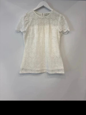 Vit spetsblus med blommönster - Söt vit blus i spets med blommigt mönster och rund hals. Blusen har korta ärmar och en feminin, lätt figurnära passform. Perfekt att styla med jeans eller kjol för en fräsch look.