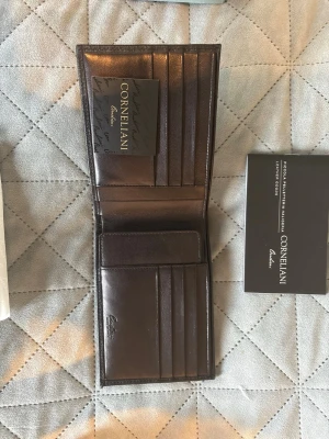 Mörkbrun plånbok från Corneliani - Snygg mörkbrun plånbok i äkta läder från Corneliani. Plånboken har flera kortfack och ett större fack för sedlar. Klassisk och stilren design Perfekt för dig som vill ha en elegant och praktisk accessoar. Klockan har aldrig använts förut. Nypris 300 euro 