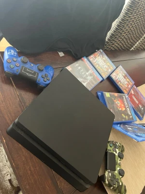 PlayStation 4 Slim med två handkontroller - Säljer en svart PlayStation 4 Slim med två DualShock 4-kontroller, en blå och en kamouflagefärgad. Flera populära spel medföljer, bland annat GTA V två stycken, Resident Evil 4, Minecraft och Until Dawn. Perfekt för gamingkvällar med vänner!