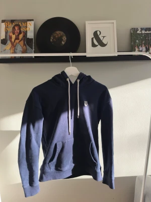 Mörkblå hoodie från maison kitsuné - Säljer en mörkblå hoodie från Maison kitsuné med klassisk huva och vita snören. Tröjan har en stor magficka och ett snyggt broderat rävmärke på bröstet. Perfekt för dig som gillar stilren design och skönt material. Skick 8/10 