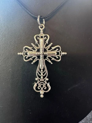 Detaljerat korshalsband i silverfärg - Säljer ett halsband med ett stort, dekorativt kors i silverfärgad metall. Korset har intrikata mönster och hänger i ett svart snöre. Perfekt för dig som gillar statement-smycken och unika detaljer.