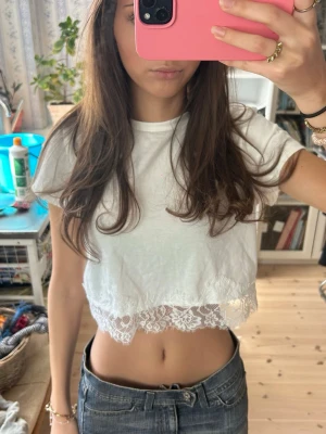 Vit croppad topp med spetskant - Söt vit croppad topp med korta ärmar och dekorativ spetskant längst ner. Toppen har en enkel, clean look med en feminin touch tack vare spetsdetaljen. Perfekt till jeans eller kjol för en trendig och avslappnad stil.