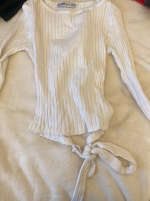 Vit ribbad långärmad topp med knyt - Säljer en vit ribbad långärmad topp med knyt i midjan. Toppen har smala vertikala ränder och är tillverkad i ett mjukt, stretchigt material. Perfekt för dig som gillar en enkel men ändå trendig stil.