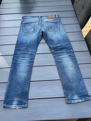 Blå raka jeans med slitningar från replay - Säljer ett par blå jeans med raka ben och tydliga slitningar samt ljusa tvättade partier. Jeansen har fem fickor, råa kanter vid bensluten och klassisk denimlook. Perfekta för en avslappnad och trendig stil. Stl w31 L 30