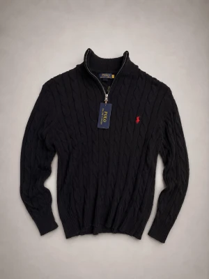 Svart kabelstickad tröja Polo Ralph Lauren - Kabelstickad svart tröja från Polo Ralph Lauren med halv dragkedja och hög krage. Tröjan har det klassiska röda broderade logot på bröstet och ribbade muddar vid ärmslut och nederkant. Perfekt för lager på lager och snygg till jeans.