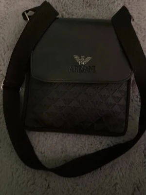 Svart axelväska från Armani - Snygg svart axelväska från Armani med justerbar axelrem. Väskan har lock med magnetknäppning, diskret mönster på framsidan och silverfärgade detaljer. Perfekt för dig som vill ha en stilren och praktisk väska till vardagen.