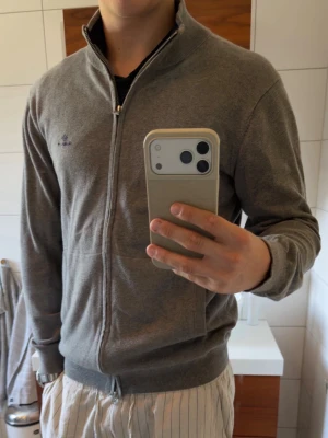 Grå ziptröja från Gant - Snygg grå ziptröja från Gant med hög krage och två fickor framtill. Tröjan har diskret logga broderad på bröstet och ribbade muddar vid ärmslut och nederkant. Perfekt att slänga på sig över en t-shirt för en clean look.