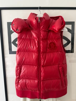 Röd dunväst med huva från Moncler - Snygg röd dunväst från Moncler med glansig finish och stor avtagbar huva. Västen har två fickor framtill, dragkedja och Moncler-logga på bröstet. Perfekt för lager på lager och är ascool