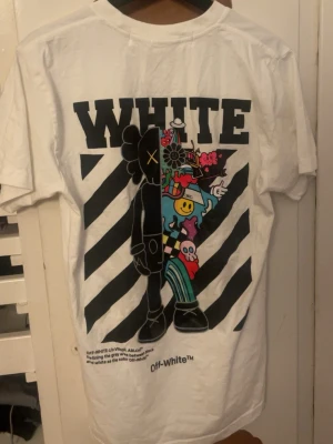 Vit Off-White t-shirt med tryck - Säljer en vit t-shirt från Off-White med stort grafiskt tryck på ryggen och mindre tryck fram. T-shirten har coola illustrationer och textdetaljer, samt klassiska svarta ränder i bakgrunden. Perfekt för dig som gillar streetwear och vill sticka ut.