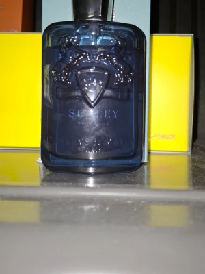 Parfym Parfums de Marly Sedley - En lyxig parfym från Parfums de Marly, modellen Sedley. Flaskan är djupblå med elegant reliefmönster och silverdetaljer. Designen är modern och exklusiv, perfekt för dig som gillar stilrena och sofistikerade dofter. Kommer i matchande blå förpackning. Själva flaskan i sig är på topp sick men det är bara kartongen till parfymen så har gått sönder lite eftersom att den är gjord av papper. Men även att den råkade få lite vatten på sig en gång. Det finns cirka 7/8 delar kvar av parfymen.