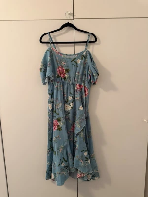 Blommig blå långklänning med volang - Söt blå långklänning med blommigt mönster och volangdetaljer. Klänningen har offshoulder-ärmar och smala axelband, vilket ger en luftig och somrig känsla. Perfekt  för varma dagar och speciella tillfällen.