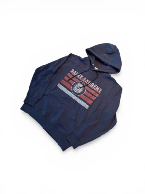 Mörkblå Saint Laurent hoodie - Snygg mörkblå hoodie från Saint Laurent med tryck i vitt och rött framtill. Klassisk huva med dragsko och stor magficka. Perfekt för dig som vill ha en stilren och trendig look. Materialet känns mjukt och bekvämt mot huden.