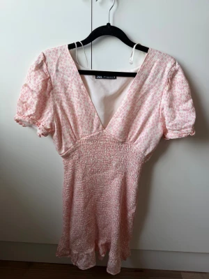 Kort blommig klänning från Zara - Söt kort klänning från Zara med ljusrosa och vita småblommor. Klänningen har v-ringning, puffärmar och smockad midja för en bekväm passform. Perfekt för sommarens varma dagar och har en lätt, luftig känsla.