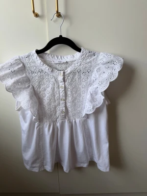 Vit broderad blus med volangärm - Supersöt vit blus med broderade detaljer och knappar framtill. Blusen har volangärmar och rund halsringning. Materialet är lätt och luftigt, perfekt för sommardagar. Blommigt hålmönster ger en romantisk vibe.
