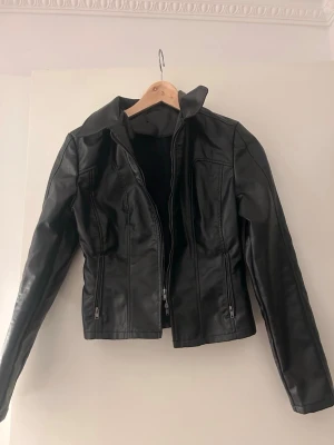 Svart skinnjacka med dragkedjor - Snygg svart skinnjacka med figurnära passform och coola detaljer som dragkedjor framtill och vid fickorna. Jackan har en klassisk krage och stilren design som passar till många olika outfits. Perfekt för dig som vill ha en tidlös och trendig look.