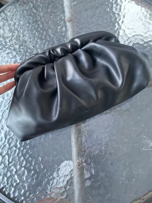 Svart kuvertväska i fejkläder - Kan fixa fler bilder ifall det behövs! Trendig svart kuvertväska i mjukt fejkläder med puffig, rynkad design. Väskan har en minimalistisk look utan synliga detaljer eller logga, perfekt att bära i handen för en stilren vibe.