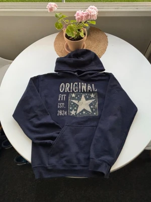 Mörkblå hoodie med stjärntryck - Mörkblå hoodie med stor huva och magficka. På bröstet finns ett tryck med texten 'ORIGINAL FIT EST. 2024' och en stor vit stjärna omgiven av mindre stjärnor. Hoodien har en avslappnad passform och ribbade muddar vid ärmar och nederkant. Storlek S men passar mindre 