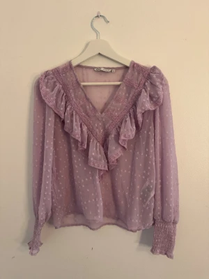 Ljuslila blus med volang och spets - Söt ljuslila blus i transparent tyg med små prickar, volangdetaljer längs V-ringningen och spetskant. Långa ärmar med smock vid handlederna ger en romantisk vibe. Perfekt för dig som gillar feminina detaljer och pastellfärger.