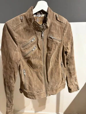 Brun mocka skinnjacka med dragkedjor - Snygg brun skinnjacka i mocka med flera silverfärgade dragkedjor framtill och coola detaljer på axlarna. Jackan har ståkrage med tryckknapp och ett mönstrat innerfoder som ger extra stilpoäng. Perfekt för dig som vill ha en trendig och unik look. Den har däremot en mörk fläck på axeln.
