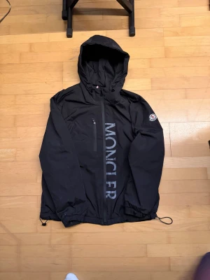 Svart vindjacka från Moncler - Snygg svart vindjacka från Moncler med stor logga längs framsidan och Moncler-märke på ärmen. Jackan har huva, dragkedja, bröstficka och justerbar nederkant med dragsko. Perfekt för dig som vill ha en stilren och sportig look.