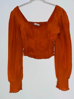 Orange långärmad blus med smock - Orange blus med långärmade, lätt puffade ärmar och smockdetaljer vid ärmslut och rygg. Blusen har en croppad passform och kan bäras både off-shoulder och inte.