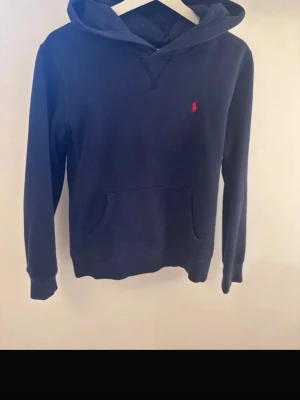 Mörkblå hoodie med röd logga - Snygg mörkblå hoodie med huva och känguruficka framtill. Hoodien har ribbade muddar vid ärmar och nederkant, och är tillverkad i mjukt material som känns skönt mot huden. Nypris:1295kr 
