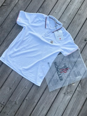 Vit Moncler pikétröja med röd detalj - Snygg vit pikétröja från Moncler med klassisk krage och knappar framtill. Tröjan har röda och blå ränder på krage och ärmslut samt Moncler-logga på bröstet. Perfekt för en clean och sportig stil.