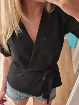 Svart omlottopp med knytband - Snygg svart omlottopp med v-ringning och knytband i midjan. Toppen har korta ärmar och en stilren, enkel design som passar till mycket. Perfekt att matcha med jeans eller kjol för en trendig look.