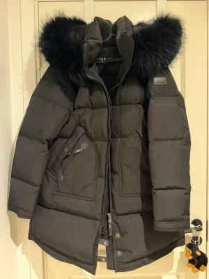 Svart vinterjacka med pälshuva Cedrico - Snygg svart dunjacka från Cedrico med stor huva och svart fuskpäls. Jackan har dragkedja framtill, två stora fickor med dragkedja och patch på ärmen. Perfekt för kalla vinterdagar och riktigt varm. Oversized passform och längre modell.